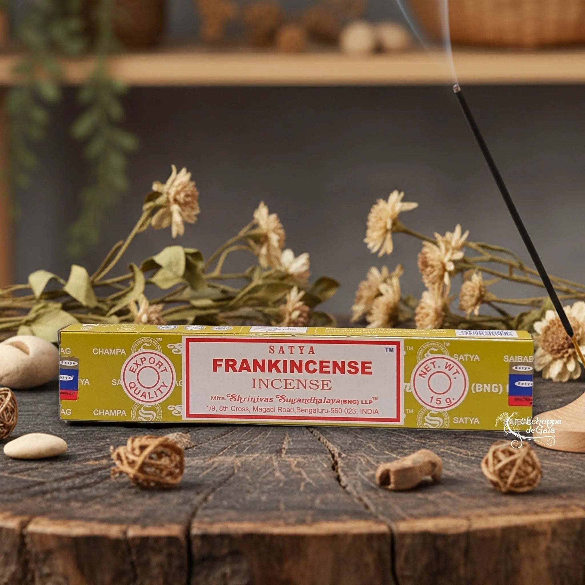 Boîte d'encens Satya "Frankincense" posée sur une table en bois avec des fleurs séchées et un bâton d'encens fumant.