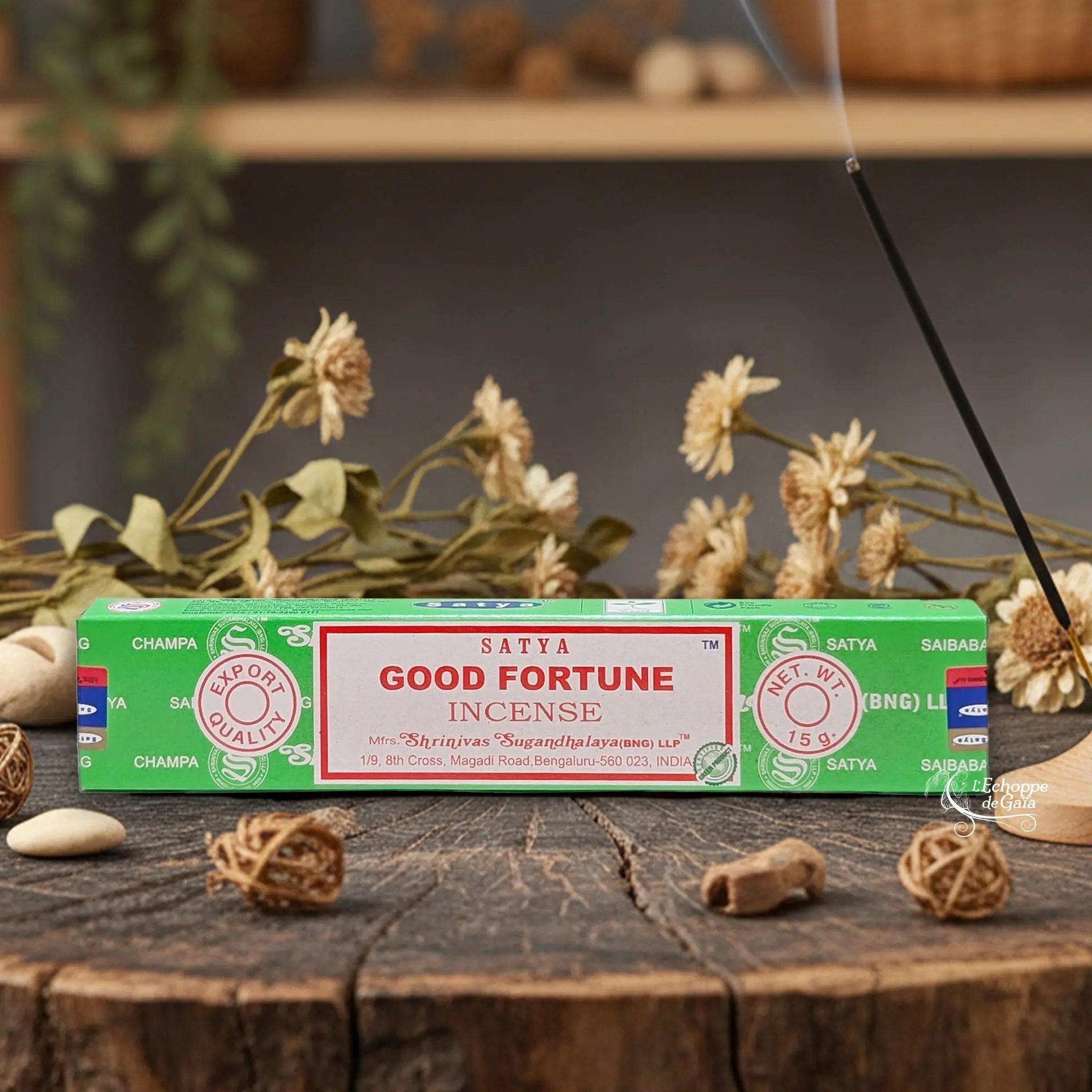 Boîte d'encens Satya "Good Fortune" posée sur une table en bois avec des fleurs séchées et un bâton d'encens fumant.