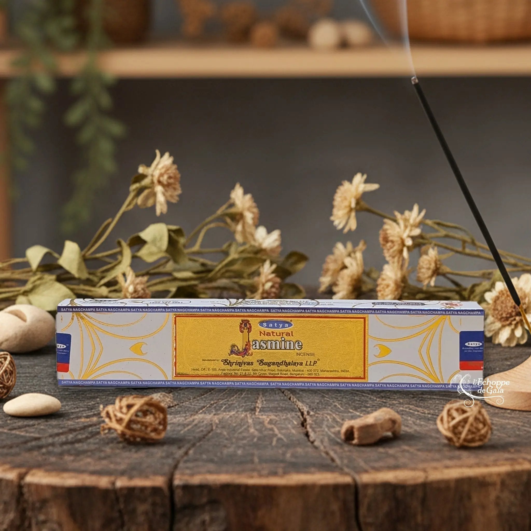 Boîte d'encens Satya "Natural Jasmine" posée sur une table en bois avec des fleurs séchées et un bâton d'encens fumant.