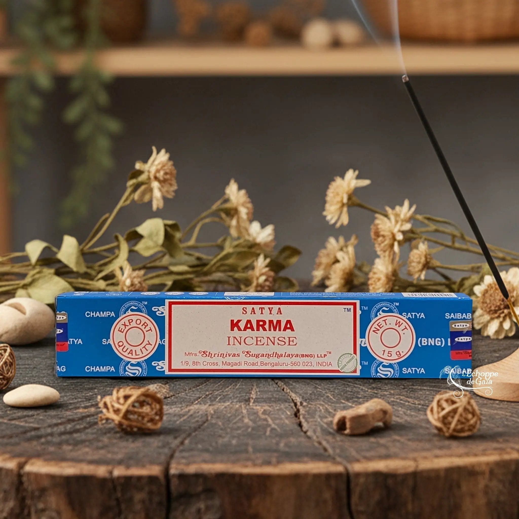 Boîte d'encens Satya "Karma" posée sur une table en bois avec des fleurs séchées et un bâton d'encens fumant.