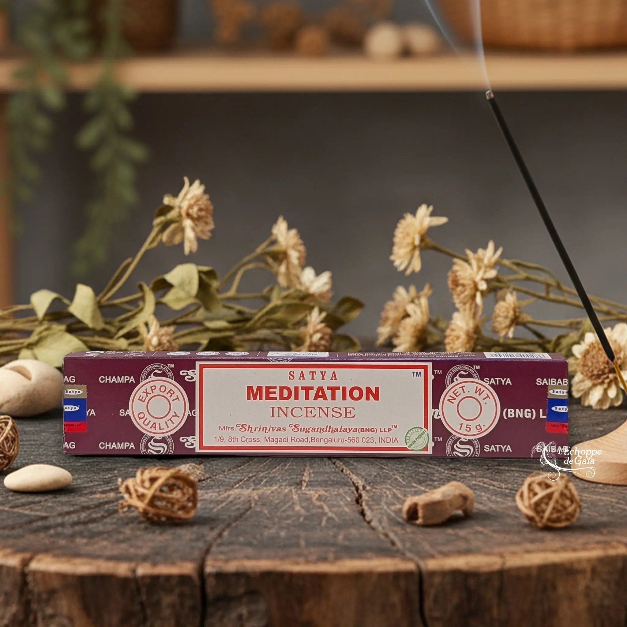 Boîte d'encens Satya "Meditation" posée sur une table en bois avec des fleurs séchées et un bâton d'encens fumant.