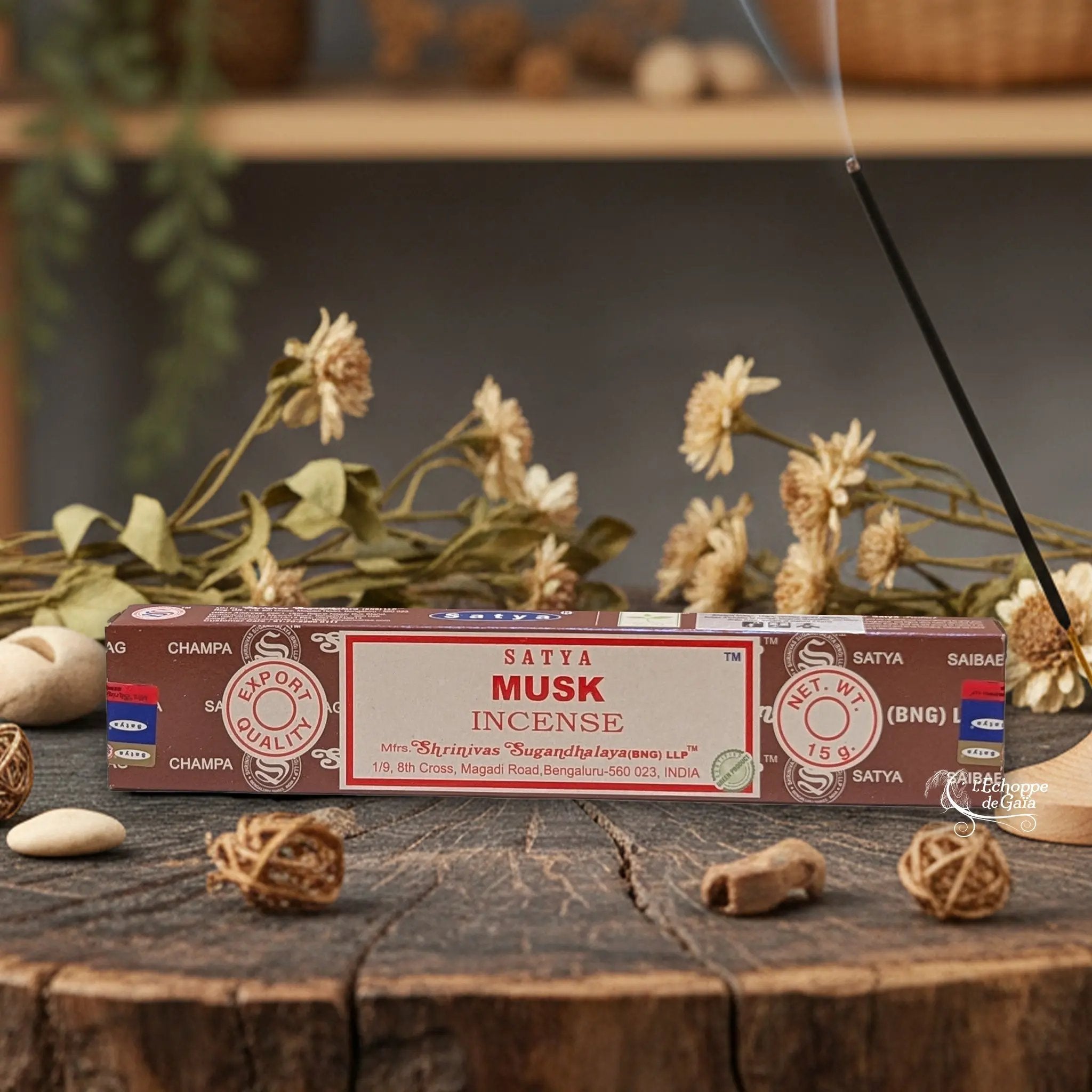 Boîte d'encens Satya "Musk" posée sur une table en bois avec des fleurs séchées et un bâton d'encens fumant.