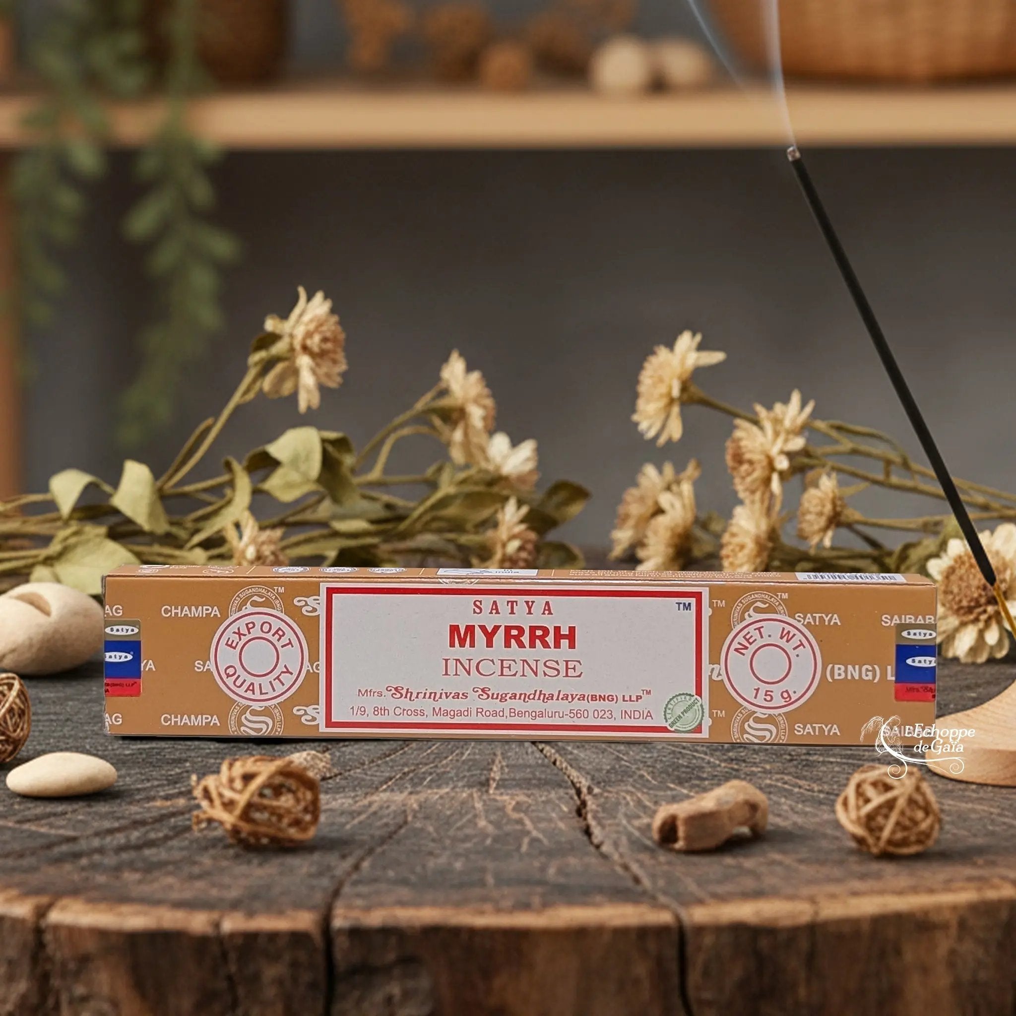 Boîte d'encens Satya "Myrrh" posée sur une table en bois avec des fleurs séchées et un bâton d'encens fumant.