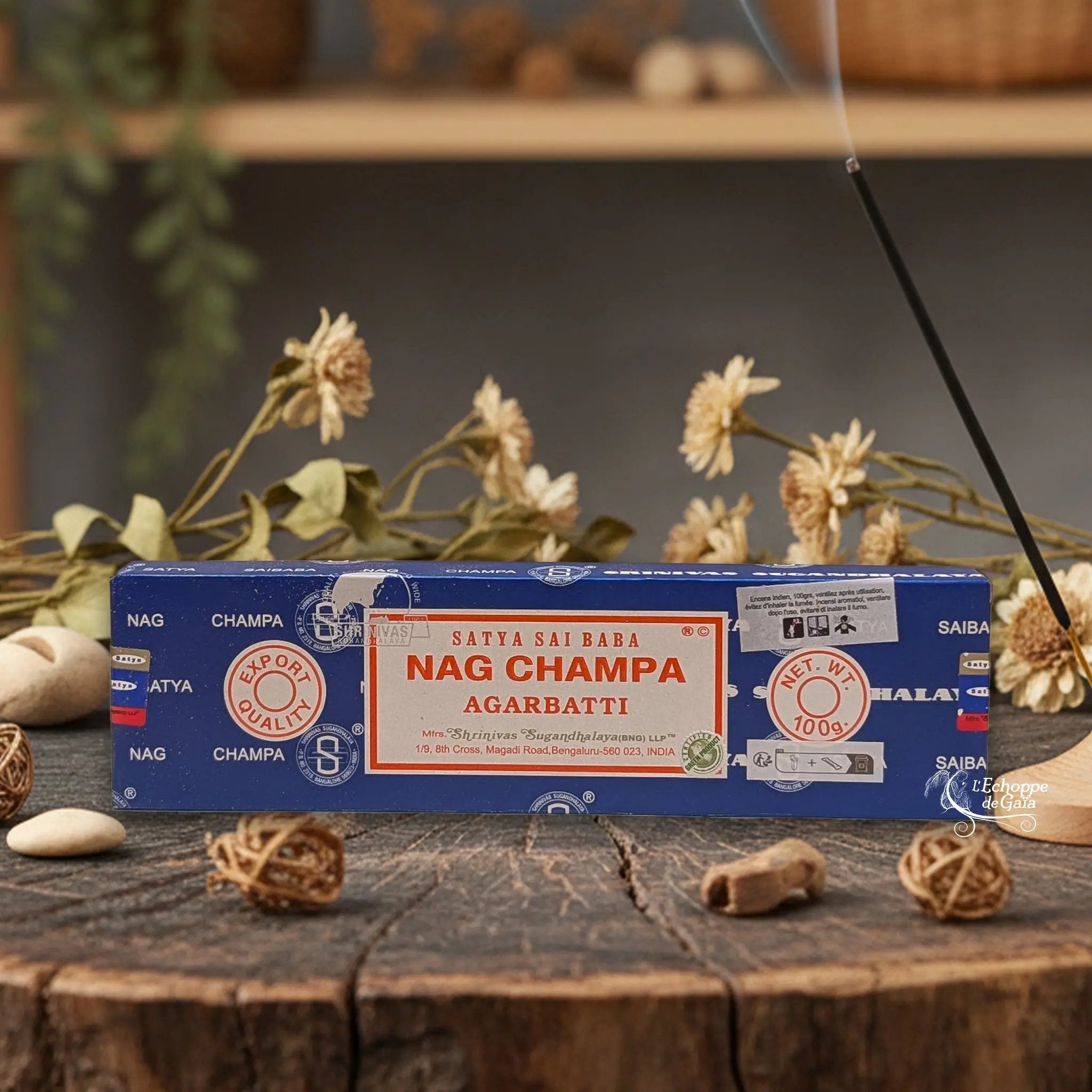 Boîte d'encens Satya Sai Baba "Nag Champa" Agarbatti posée sur une table en bois avec des fleurs séchées et un bâton d'encens fumant.
