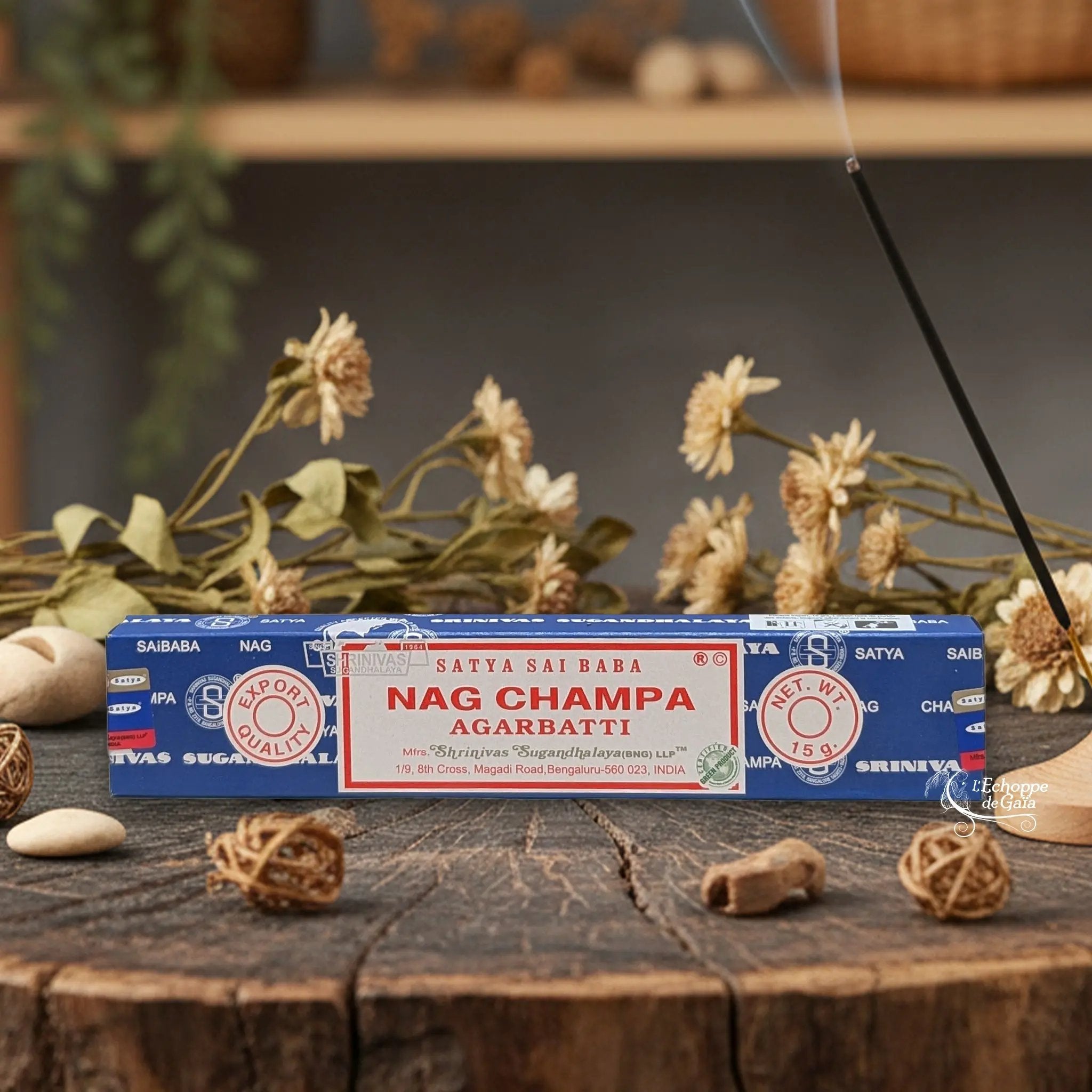 Boîte d'encens Satya Sai Baba "Nag Champa" Agarbatti posée sur une table en bois avec des fleurs séchées et un bâton d'encens fumant.
