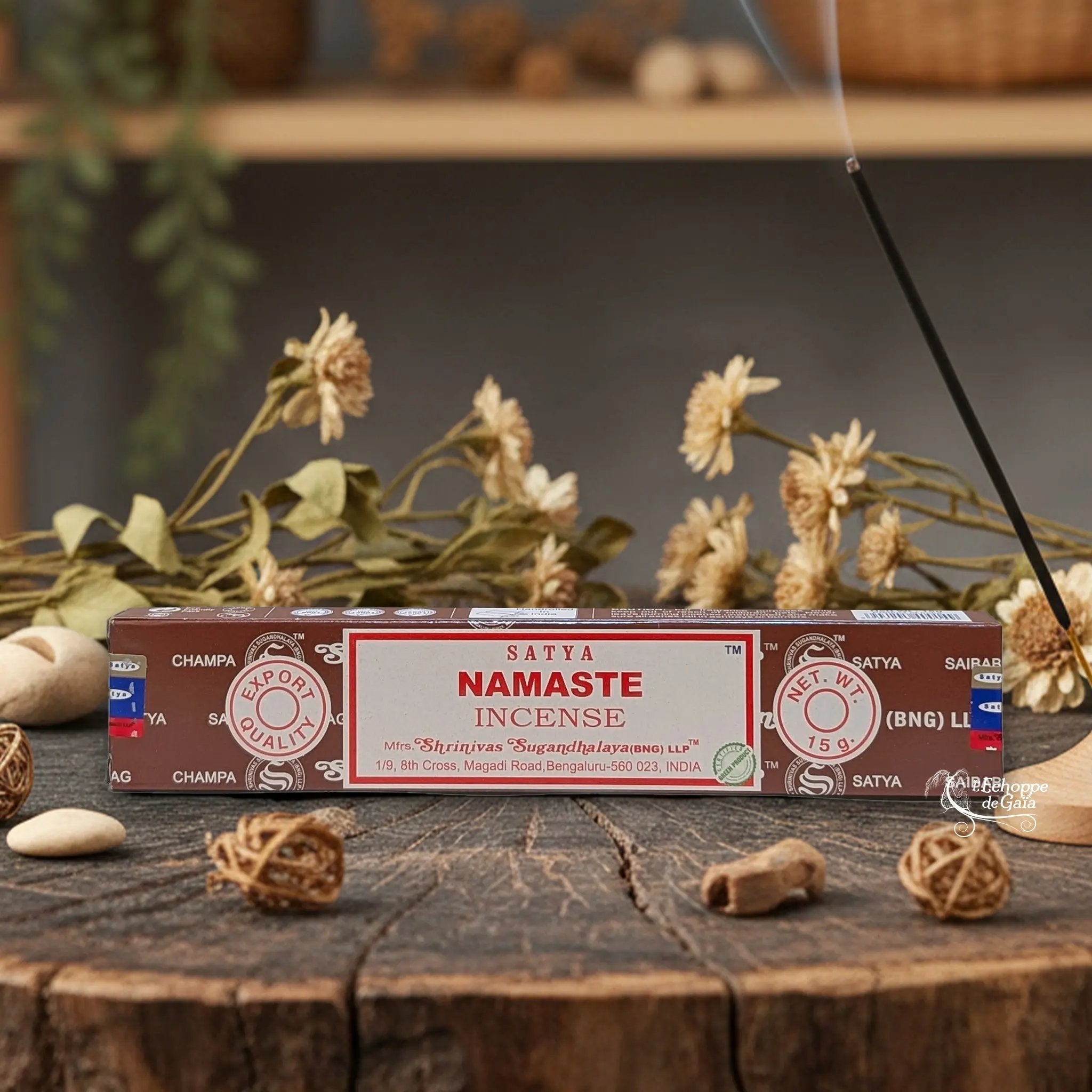 Boîte d'encens Satya "Namaste" posée sur une table en bois avec des fleurs séchées et un bâton d'encens fumant.