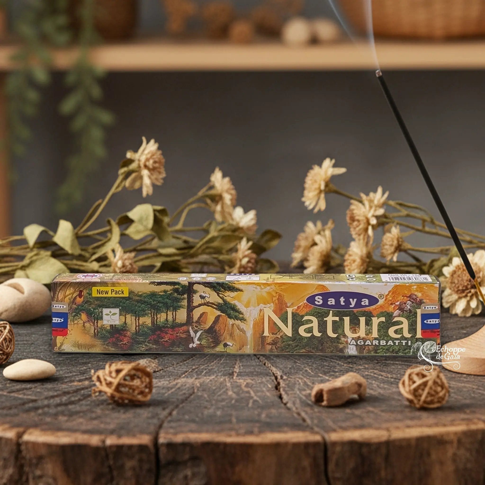 Boîte d'encens Satya "Natural" Agarbatti posée sur une table en bois avec des fleurs séchées et un bâton d'encens fumant.