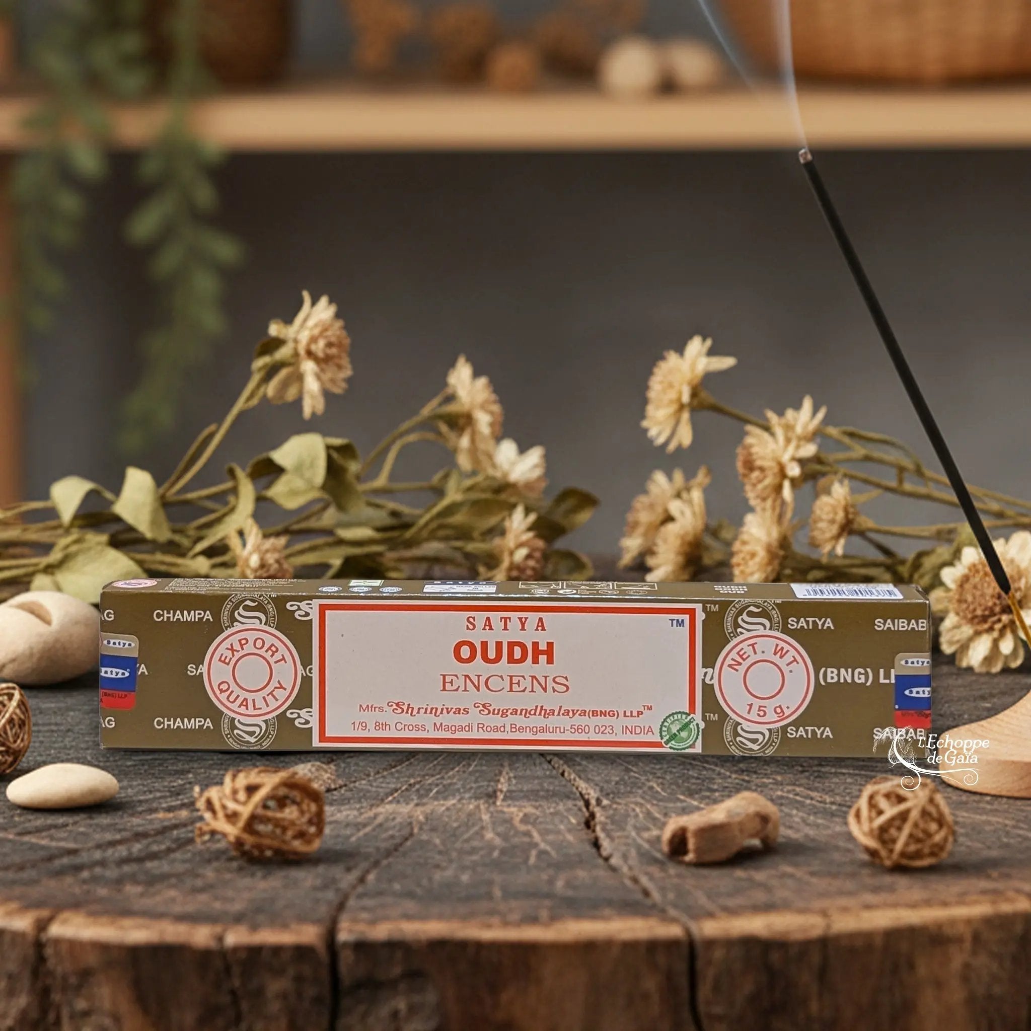 Boîte d'encens Satya "Oudh" posée sur une table en bois avec des fleurs séchées et un bâton d'encens fumant.