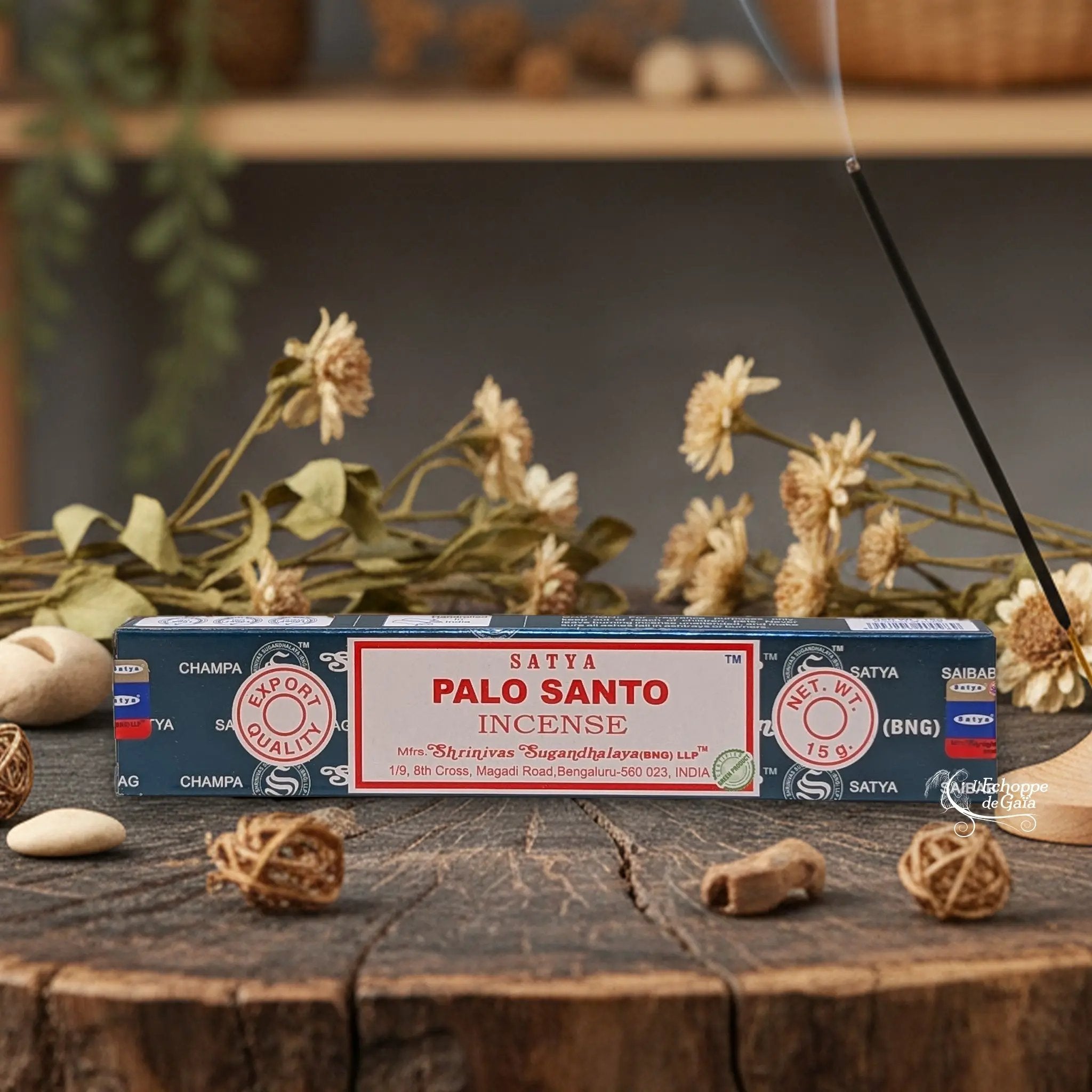 Boîte d'encens Satya "Palo Santo" posée sur une table en bois avec des fleurs séchées et un bâton d'encens fumant.