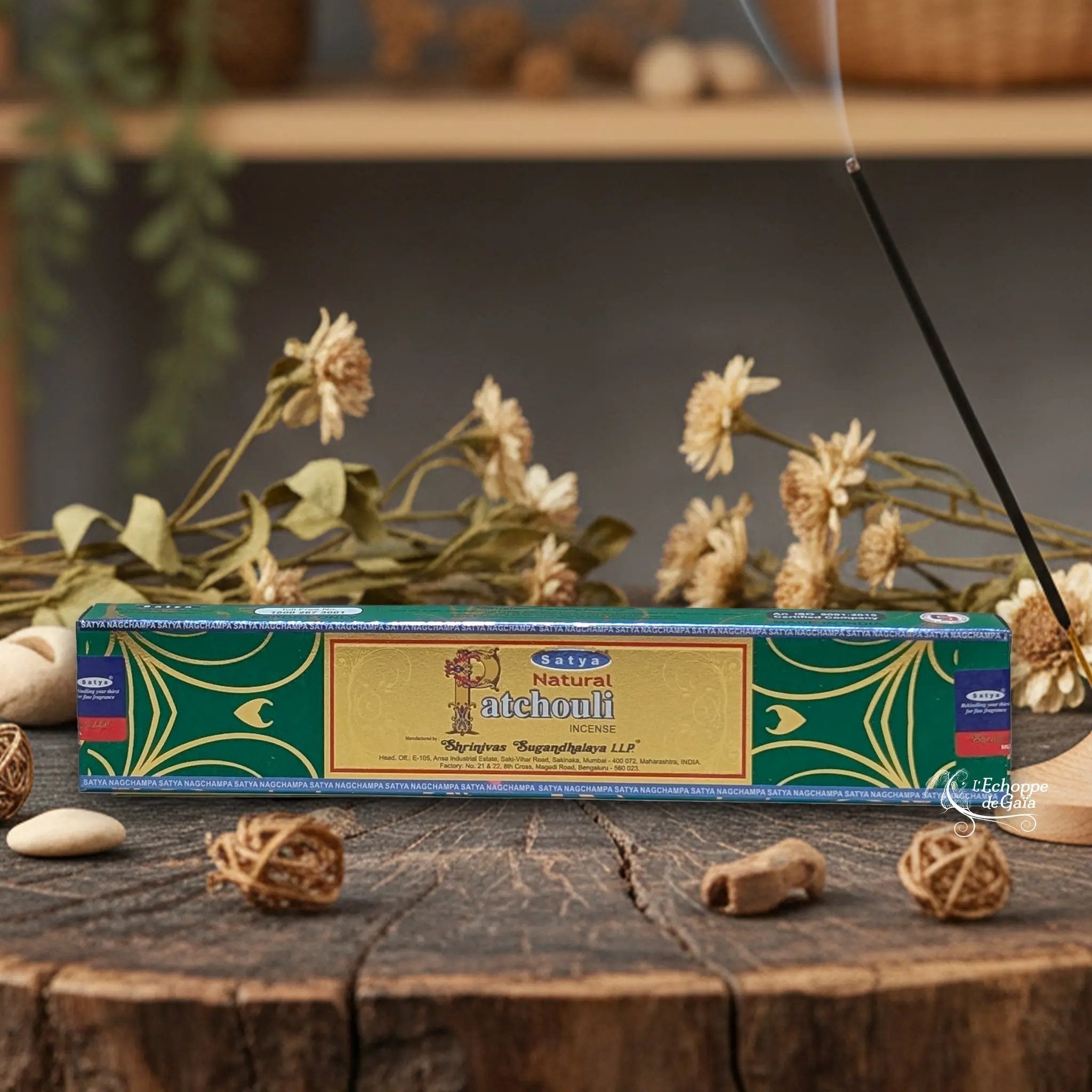 Boîte d'encens Satya "Natural Patchouli" posée sur une table en bois avec des fleurs séchées et un bâton d'encens fumant.