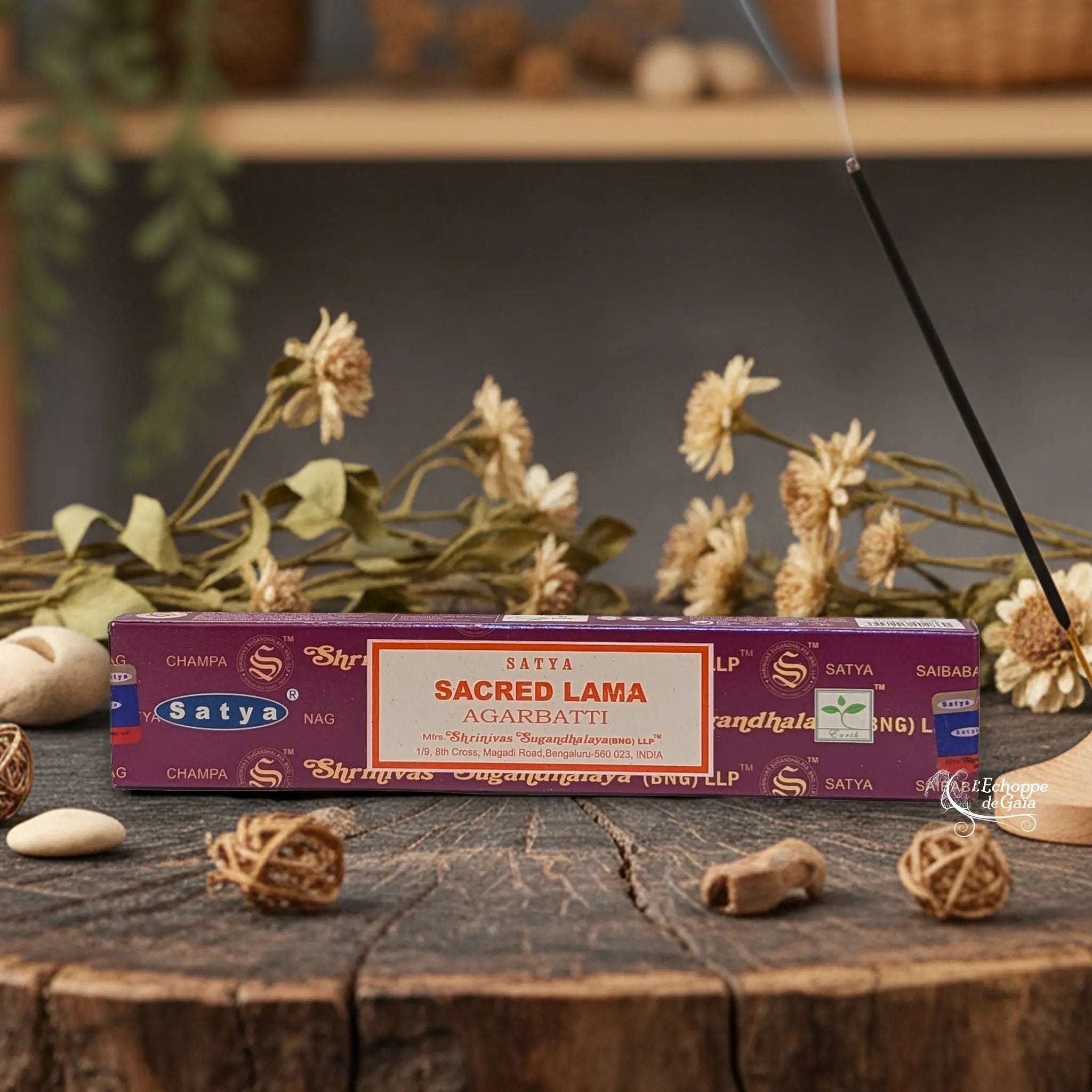 Boîte d'encens Satya "Sacred Lama" Agarbatti posée sur une table en bois avec des fleurs séchées et un bâton d'encens fumant.