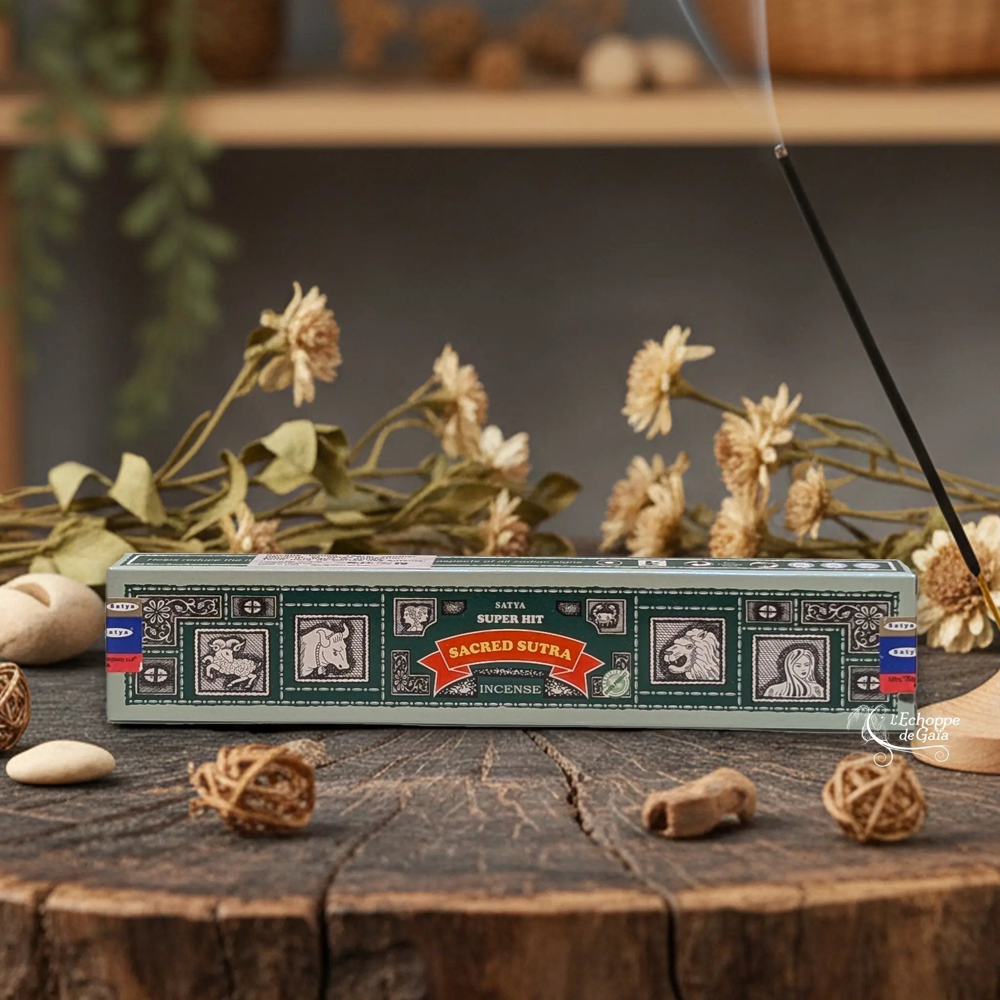 Boîte d'encens Satya Super Hit "Sacred Sutra" posée sur une table en bois avec des fleurs séchées et un bâton d'encens fumant.