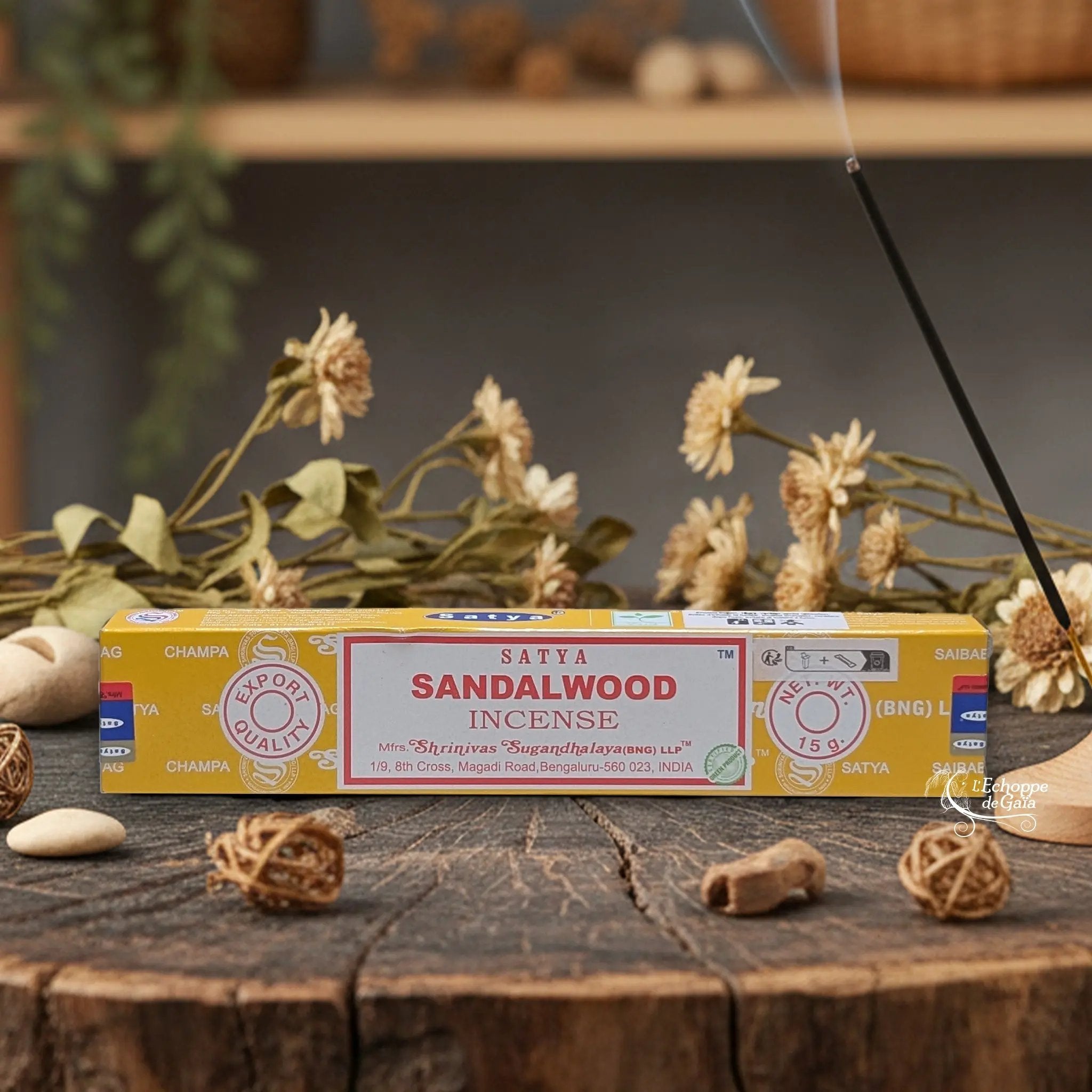 Boîte d'encens Satya "Sandalwood" posée sur une table en bois avec des fleurs séchées et un bâton d'encens fumant.