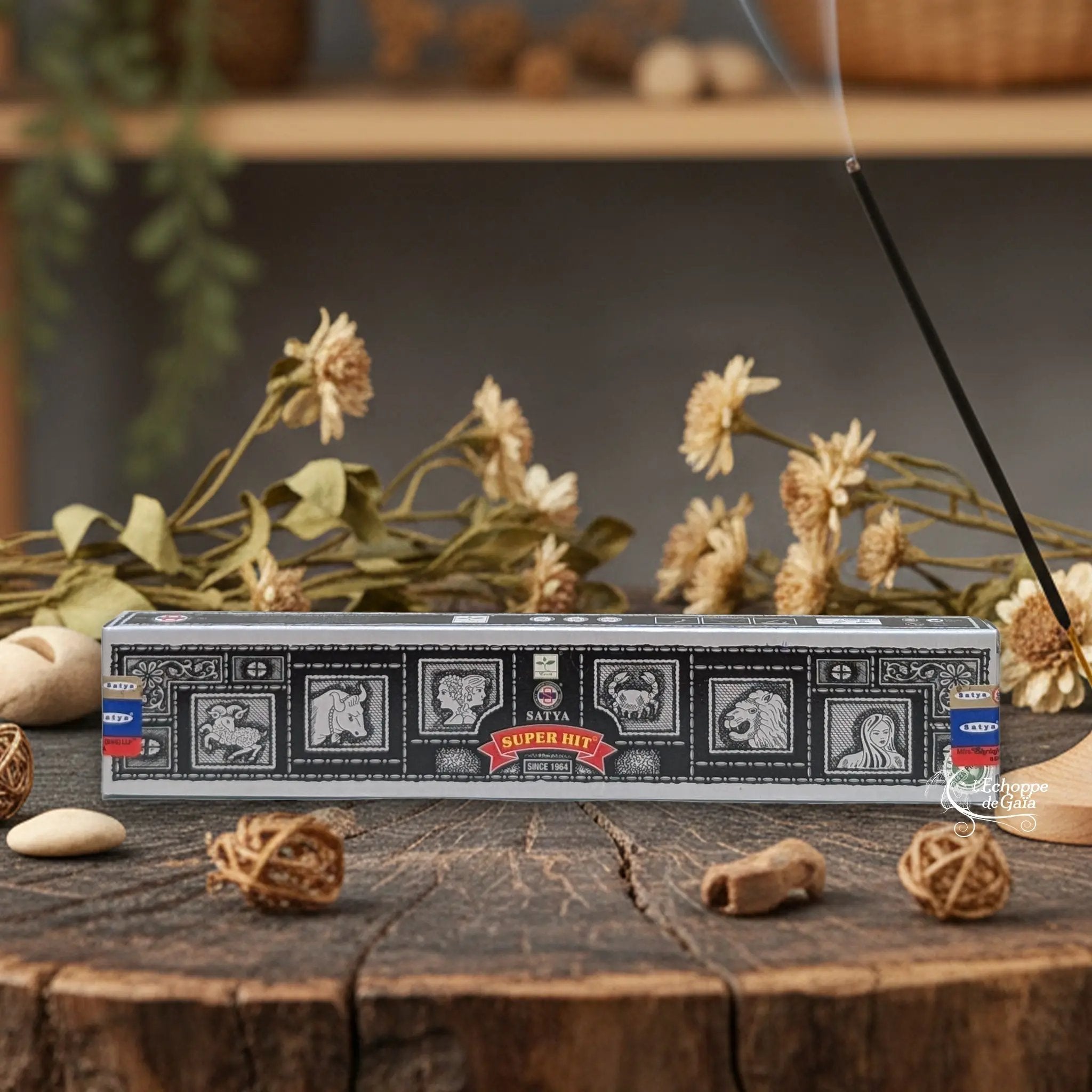 Boîte d'encens Satya "Super Hit" posée sur une table en bois avec des fleurs séchées et un bâton d'encens fumant.