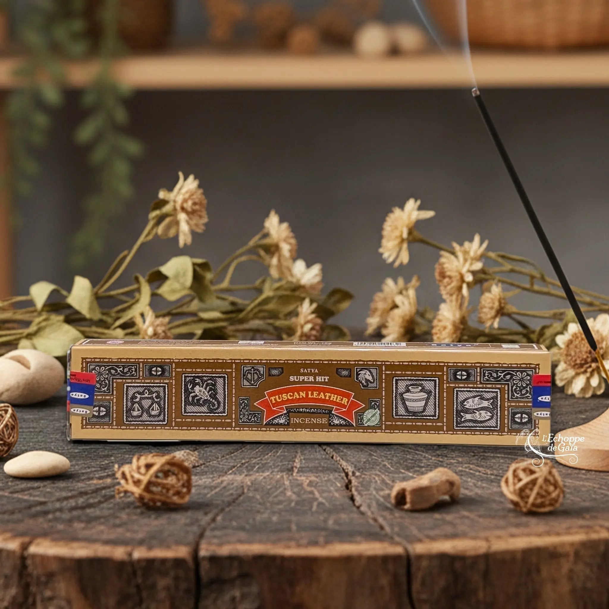 Boîte d'encens Satya Super Hit "Tuscan Leather" posée sur une table en bois avec des fleurs séchées et un bâton d'encens fumant.