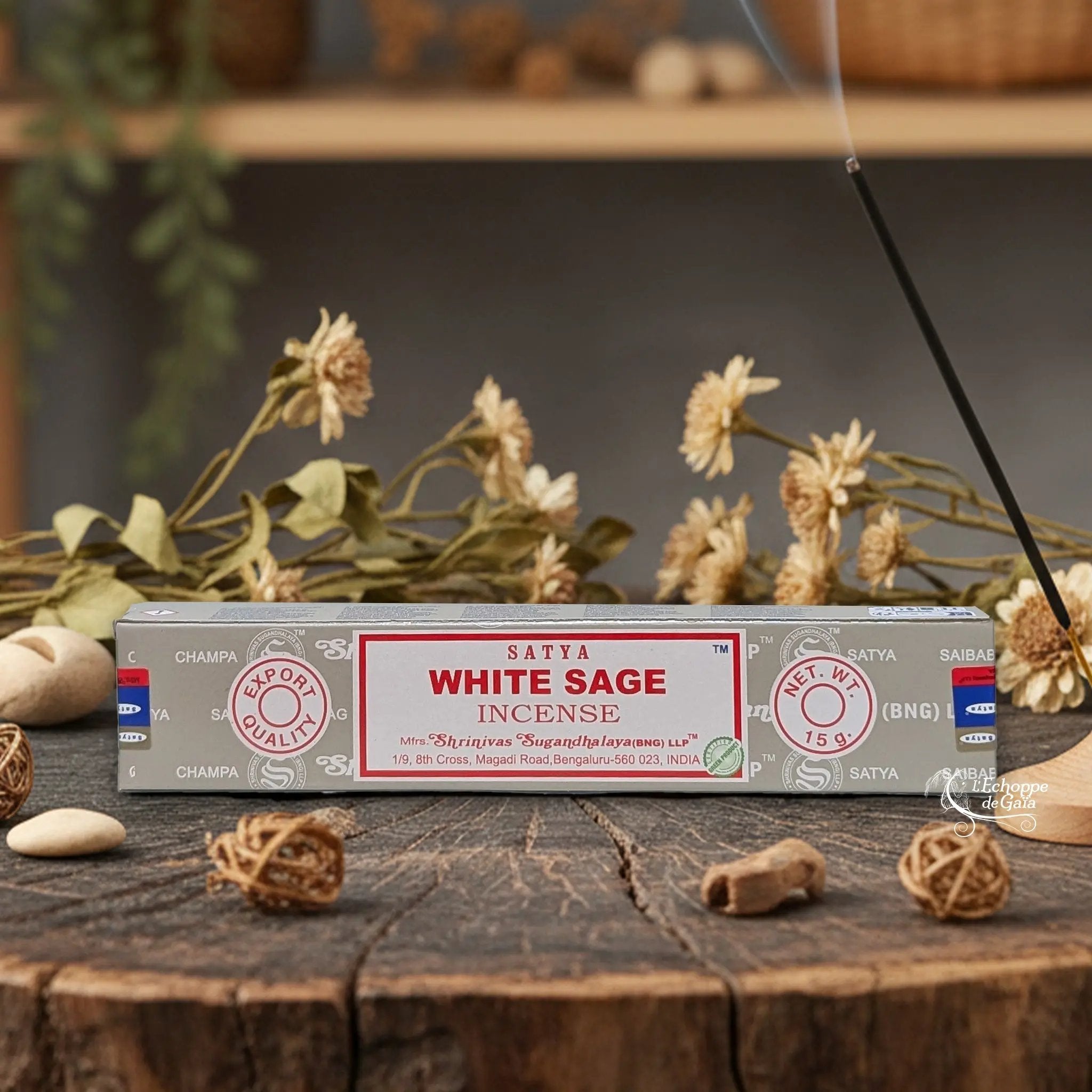Boîte d'encens Satya "White Sage" posée sur une table en bois avec des fleurs séchées et un bâton d'encens fumant.