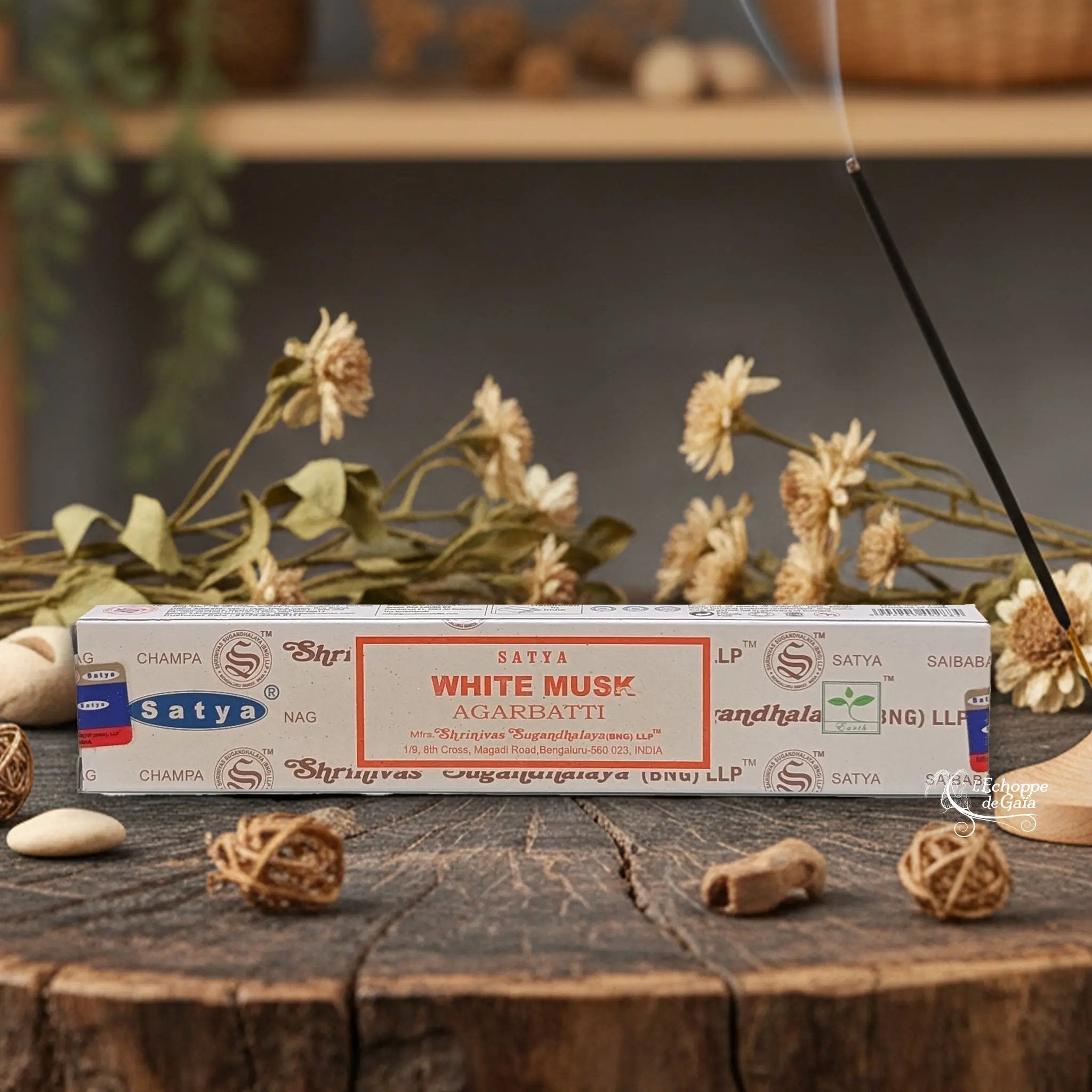 Boîte d'encens Satya "White Musk" Agarbatti posée sur une table en bois avec des fleurs séchées et un bâton d'encens fumant.