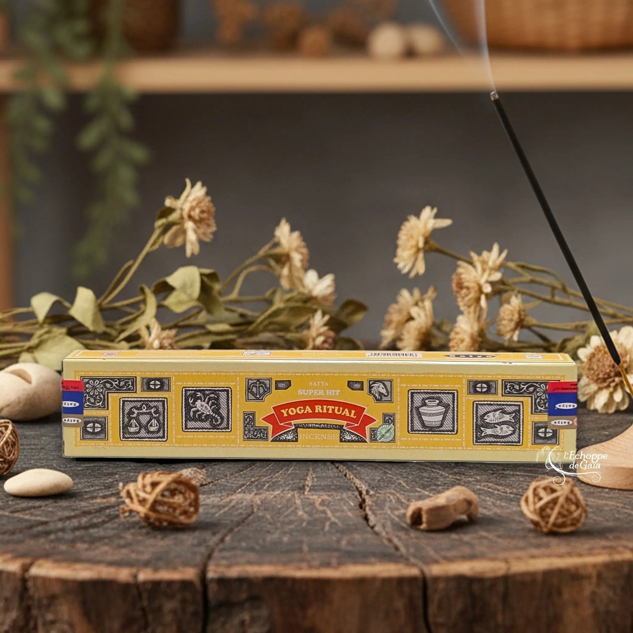 Boîte d'encens Satya Super Hit "Yoga Ritual" posée sur une table en bois avec des fleurs séchées et un bâton d'encens fumant.