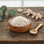 Grains de Sel Rose de l'Himalaya (couleur rose pâle à blanc), dans un petit bol en bois clair avec une cuillère, sur une planche de bois rustique.