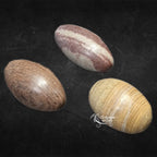 Trois pierres de shiva lingam polies de forme ovoïde, présentant des teintes beiges et des marques brunes circulaires ou rubanées, présentées sur un fond noir.