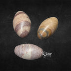 Trois pierres de shiva lingam polies de forme ovoïde, présentant des teintes beiges et des marques brunes circulaires ou rubanées, présentées sur un fond noir.