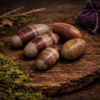 Pierres sacrées Shiva Lingam de forme ovoïde aux nuances terreuses et bandes brunes, disposées sur un plateau de bois ancien dans une ambiance de rituel spirituel.