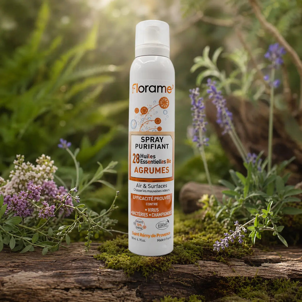 Flacon de spray purifiant bio Florame aux agrumes posé sur une écorce moussue, entouré de fleurs sauvages violettes et blanches dans une lumière naturelle.