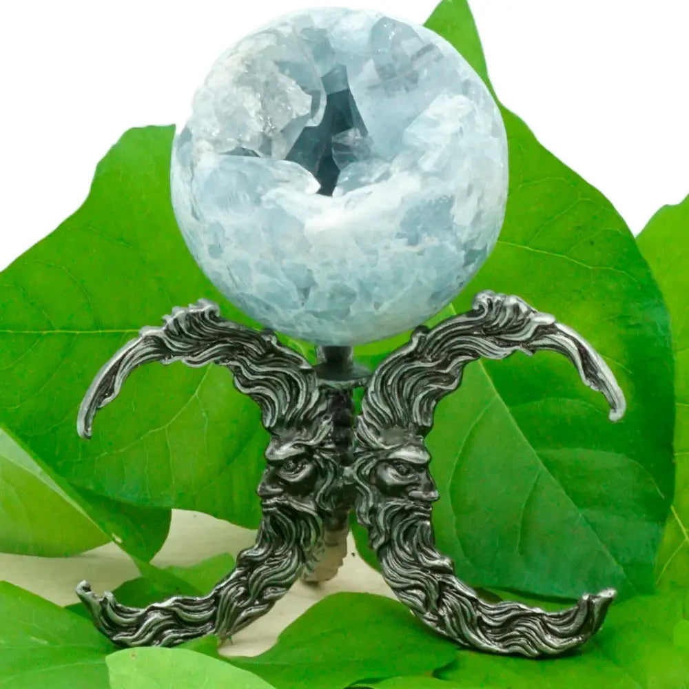 Support pour boule de cristal en métal gris argenté (aspect vieilli), motif “double croissant” symétrique composé de grandes volutes façon mèches/flammèches. On distingue deux visages barbus de profil intégrés au décor, se faisant face au centre. Une boule minérale translucide bleu pâle (type cristal/pierre) est posée au sommet sur un petit axe central. Mise en scène sur fond de grandes feuilles vertes.
