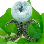 Support pour boule de cristal en métal gris argenté (aspect vieilli), motif “double croissant” symétrique composé de grandes volutes façon mèches/flammèches. On distingue deux visages barbus de profil intégrés au décor, se faisant face au centre. Une boule minérale translucide bleu pâle (type cristal/pierre) est posée au sommet sur un petit axe central. Mise en scène sur fond de grandes feuilles vertes.