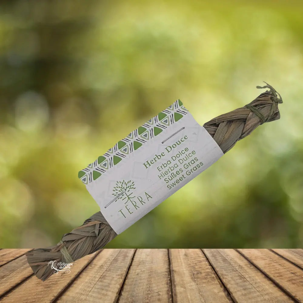 Tresse d’herbe douce (sweet grass) de la marque Terra, posée sur une table en bois devant un fond vert flou. La tresse est enveloppée d’un bandeau cartonné indiquant « Herbe Douce / Sweet Grass » en plusieurs langues, présentation type packshot pour boutique ésotérique ou bien-être.