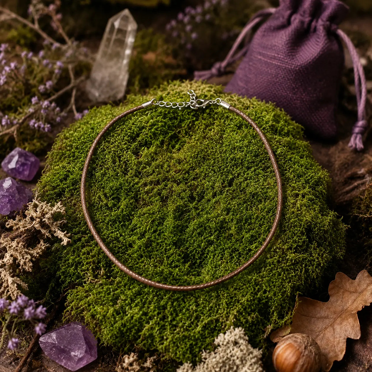 Cordon de tour de cou marron avec fermoir argenté disposé en cercle sur un lit de mousse verte, entouré de cristaux de quartz, de pierres violettes, d'une bourse en tissu violet et d'éléments naturels de sous-bois.
