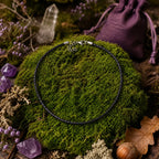 Cordon de tour de cou noir tressé avec un fermoir argenté, disposé en cercle sur un monticule de mousse verte et entouré de cristaux de quartz, de pierres violettes, d'une petite bourse en tissu violet et d'éléments de sous-bois.