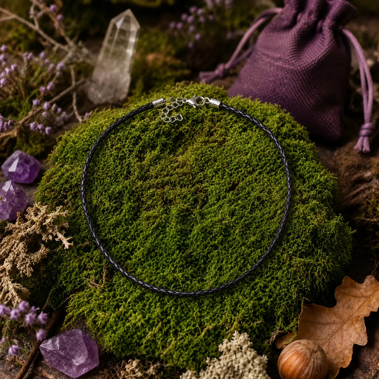Cordon de tour de cou noir tressé avec un fermoir argenté, disposé en cercle sur un monticule de mousse verte et entouré de cristaux de quartz, de pierres violettes, d'une petite bourse en tissu violet et d'éléments de sous-bois.