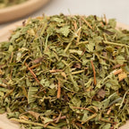 Gros plan sur un tas de verveine citronnée séchée, mélange de feuilles et petites tiges vert clair à brun, posé sur une assiette en bois claire.