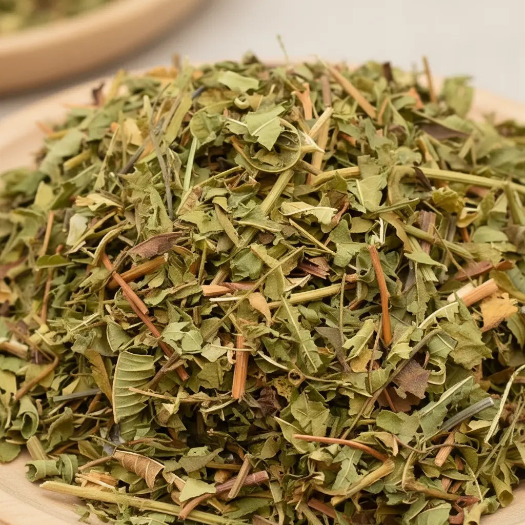 Gros plan sur un tas de verveine citronnée séchée, mélange de feuilles et petites tiges vert clair à brun, posé sur une assiette en bois claire.