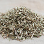 Gros plan sur un petit tas de verveine séchée, composé de fins fragments de tiges et de feuilles vert-brun sur une toile beige.